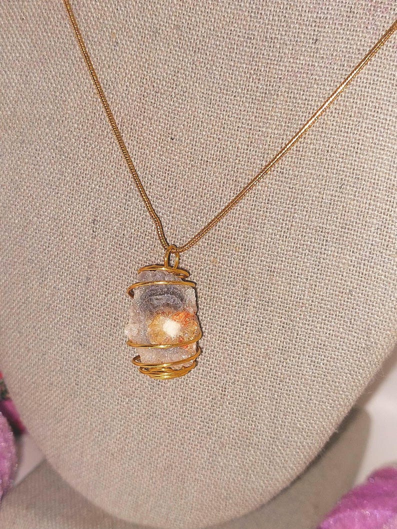 Puede incluir: Un collar con colgante envuelto en alambre dorado. El colgante presenta una piedra de color claro con inclusiones naranjas y blancas. La cadena del collar es dorada y el fondo es de tela neutra.