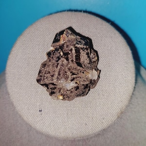 May include: A dark brown, faceted crystal with a rough, textured surface. The crystal is displayed on a light beige fabric background.