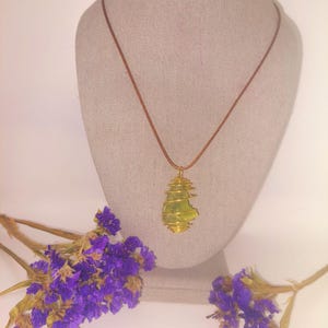 May include: A handmade necklace featuring a light green stone wrapped in gold-colored wire, suspended from a brown cord. The necklace is displayed on a mannequin bust. Purple dried flowers are arranged around the bust.