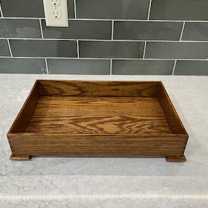 May include: A rectangular wooden tray with a natural wood grain finish. The tray has a raised edge and sits on four small feet. The wood has a warm, brown tone, and the tray appears to be handmade.