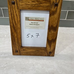 May include: A wooden picture frame with a natural wood grain finish. The frame is rectangular with a 5x7 inch opening. The frame has a label that says "Foggy Ridge Fine Handcrafted Items" and "Ray Giraud Maker/Designer".