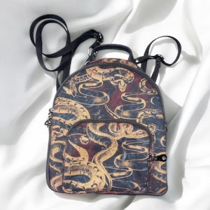 Könnte beinhalten: Ein kleiner Rucksack mit schwarzem Reißverschluss und Trägern. Der Rucksack hat ein sich wiederholendes Muster aus goldenen Schlangen und Totenköpfen auf einem dunkelblauen und burgunderroten Hintergrund. Die Tasche hat eine kleine Vordertasche und einen oberen Griff.