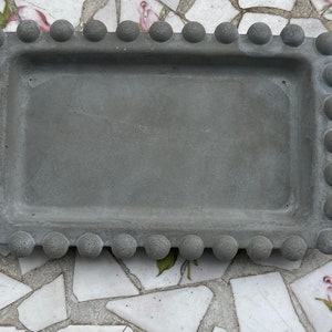 May include: A grey concrete rectangular tray with a beaded border. The tray is sitting on a white and grey tiled surface.