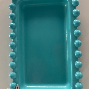 May include: A light blue rectangular tray with a beaded edge. The tray has a decorative silver flower design on the bottom left corner.
