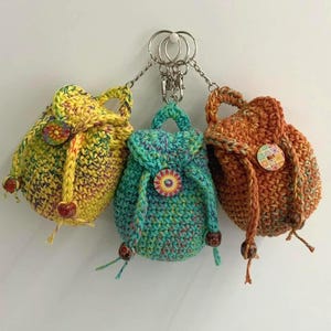 May include: Three miniature crochet backpack keychains in yellow, teal, and orange. Each bag features a drawstring closure, a top handle, and a decorative circular button on the front flap. The bags are attached to silver-toned metal key rings.