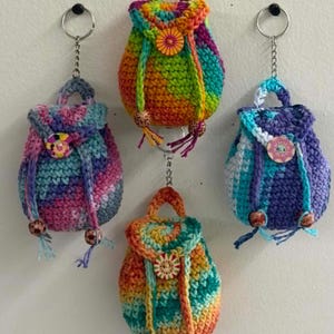 May include: Four miniature crochet backpacks hanging from metal keychains. Each bag features variegated yarn in bright colors, a top handle, a flap secured with a decorative button, and two drawstring cords ending in small wooden beads at the bottom.