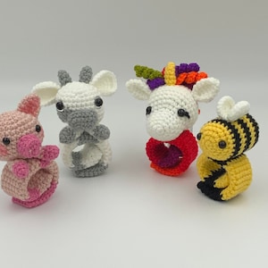 May include: Four crocheted animal napkin rings: a pink pig, a white and gray cow, a white unicorn with a rainbow mane, and a yellow and black bee. Each ring is designed to hold a napkin and features detailed facial features.