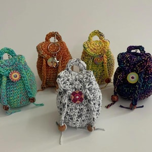 May include: A collection of five crocheted mini backpacks in various colors: teal, orange, yellow, gray, and purple. Each backpack features a drawstring closure, a decorative button, and wooden bead accents. Some have keychains attached.