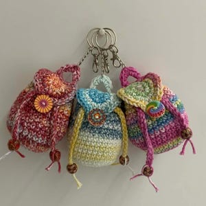 May include: Three handmade crochet backpack keychains in shades of red, blue, and pink. Each mini backpack features a button closure and wooden bead accents. The keychains are attached to silver metal rings.