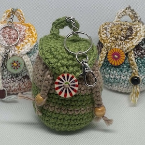 May include: Three crocheted backpack keychains in different colors and patterns. The keychains are made with a variety of yarn colors, including green, brown, beige, white, and blue. Each keychain features a small button with a colorful design.