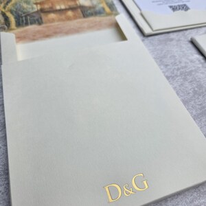 K&ouml;nnte beinhalten: Elfenbeinfarbene Hochzeitseinladung mit Landschaftsmalerei und goldfarbenen Buchstaben. Die Suite enth&auml;lt eine Einladung, eine Antwortkarte und eine Tasche mit den goldenen Buchstaben "D&G".