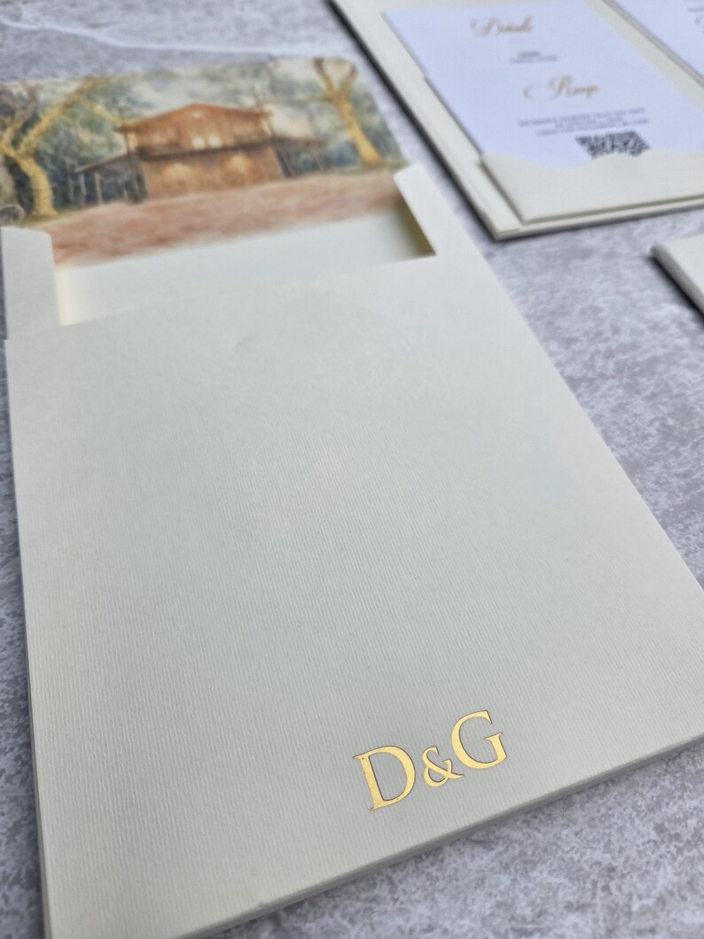K&ouml;nnte beinhalten: Elfenbeinfarbene Hochzeitseinladung mit Landschaftsmalerei und goldfarbenen Buchstaben. Die Suite enth&auml;lt eine Einladung, eine Antwortkarte und eine Tasche mit den goldenen Buchstaben "D&G".