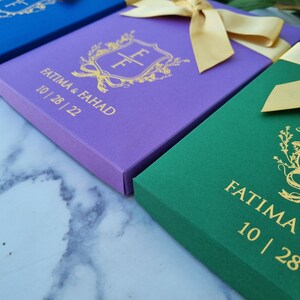 May include: Three rectangular boxes in blue, purple, and green, each topped with a gold satin ribbon. The boxes feature a gold foil crest with the initials FF, the names FATIMA & FAHAD, and the date 10 | 28 | 22 printed on the front surface.