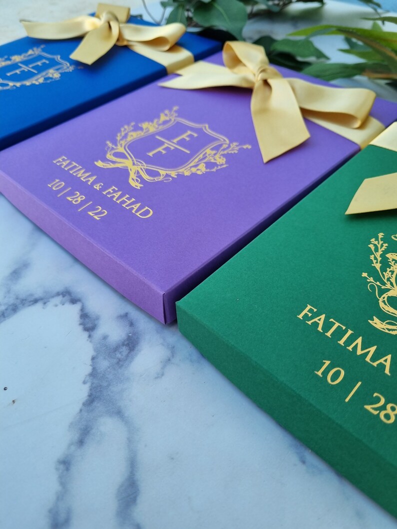 May include: Three rectangular boxes in blue, purple, and green, each topped with a gold satin ribbon. The boxes feature a gold foil crest with the initials FF, the names FATIMA & FAHAD, and the date 10 | 28 | 22 printed on the front surface.