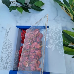 May include: A clear acrylic wedding invitation with gold lettering, featuring the names Fatima Shaikh and Muhammad Fahad Nasir. The invitation is placed over a blue box filled with red dried flower petals, flanked by two translucent white vellum panels.