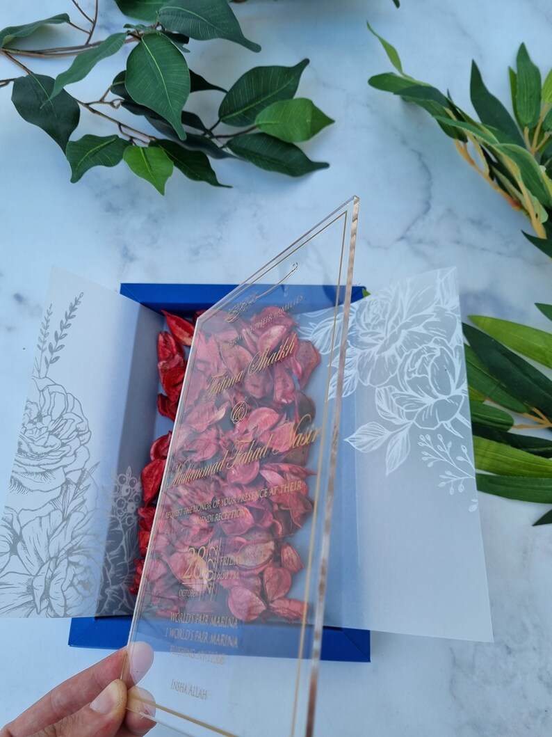May include: A clear acrylic wedding invitation with gold lettering, featuring the names Fatima Shaikh and Muhammad Fahad Nasir. The invitation is placed over a blue box filled with red dried flower petals, flanked by two translucent white vellum panels.