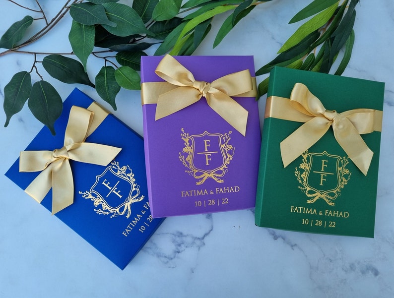 May include: Three rectangular gift boxes in royal blue, purple, and dark green, each adorned with a gold satin ribbon bow. The front of each box features a gold foil crest with the letters FF, the names FATIMA & FAHAD, and the date 10 | 28 | 22.