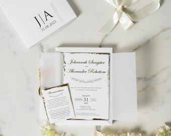 May include: A white wedding invitation set featuring a gatefold card with gold-edged paper, printed with the names Johannah Sangster and Alexander Robottom, an accommodation card, and a matching white box with the initials J|A and date 31.08.2023.