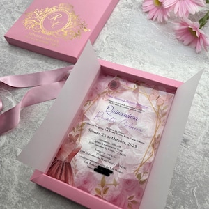 May include: A pink invitation box with gold lettering and a matching lid. The box contains an invitation with the name "Renata Cabrera" and the date "October 25, 2025". Pink gerbera daisies and a pink ribbon are also visible.