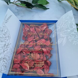 May include: A rectangular wedding invitation featuring a clear acrylic panel filled with dried red flower petals. The panel is encased in a blue folder with white floral line art on the inner flaps. Gold text displays the names Fatima Shaikh and Muhammad Fahad Nasir, the date October 28, 2022, and the venue location at World's Fair Marina in Flushing, NY.