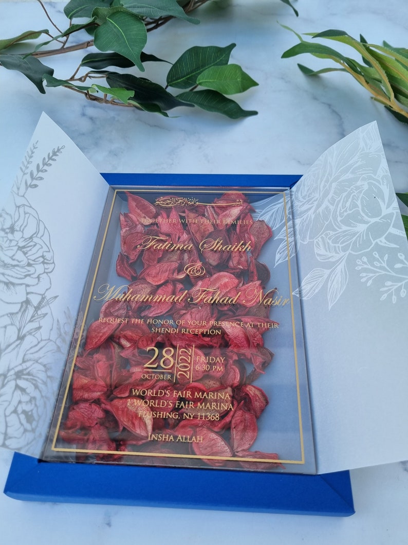 May include: A rectangular wedding invitation featuring a clear acrylic panel filled with dried red flower petals. The panel is encased in a blue folder with white floral line art on the inner flaps. Gold text displays the names Fatima Shaikh and Muhammad Fahad Nasir, the date October 28, 2022, and the venue location at World's Fair Marina in Flushing, NY.