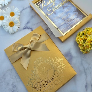 Invitation de mariage luxe avec boîte or perlé, noms en acrylique miroir or et pétales blancs.