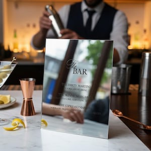 May include: A mirrored sign with the text "The BAR" and a list of cocktails, including "The Dirty Martini" and "The Skinny Margarita." A martini glass with olives, a copper jigger, and a spoon are on a marble countertop.