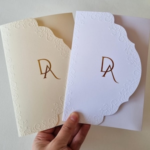 Puede incluir: Dos sobres de invitación de boda blancos con un borde decorativo festoneado. Los sobres están en relieve con un patrón floral. Los sobres están en blanco excepto por las iniciales "DA" en lámina dorada.