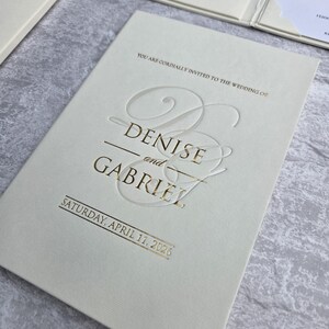 K&ouml;nnte beinhalten: Elfenbeinfarbene Hochzeitseinladung mit goldfarbenem Folienschriftzug. Die Einladung lautet "Denise und Gabriel" und "Samstag, 11. April 2026". Die Einladung ist Teil eines Sets mit einer Tasche auf der rechten Seite.