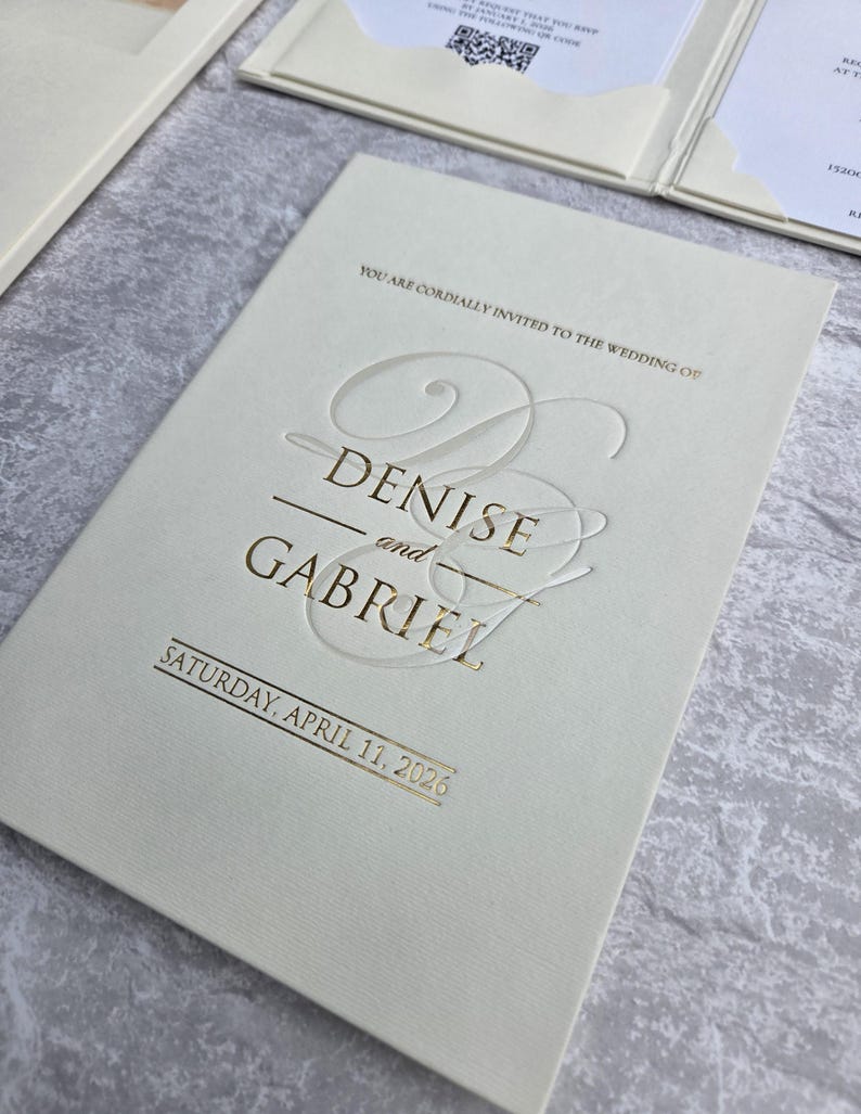 K&ouml;nnte beinhalten: Elfenbeinfarbene Hochzeitseinladung mit goldfarbenem Folienschriftzug. Die Einladung lautet "Denise und Gabriel" und "Samstag, 11. April 2026". Die Einladung ist Teil eines Sets mit einer Tasche auf der rechten Seite.