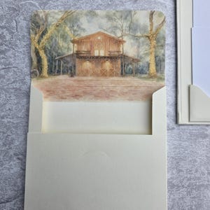 K&ouml;nnte beinhalten: Eine cremefarbene Einladungskarte mit einem Aquarellgem&auml;lde einer rustikalen Scheune mit Lichterketten. Die Karte wird in einen cremefarbenen Umschlag gesteckt, mit einer passenden Karte auf der rechten Seite. Der Hintergrund ist eine strukturierte, hellgraue Oberfl&auml;che.
