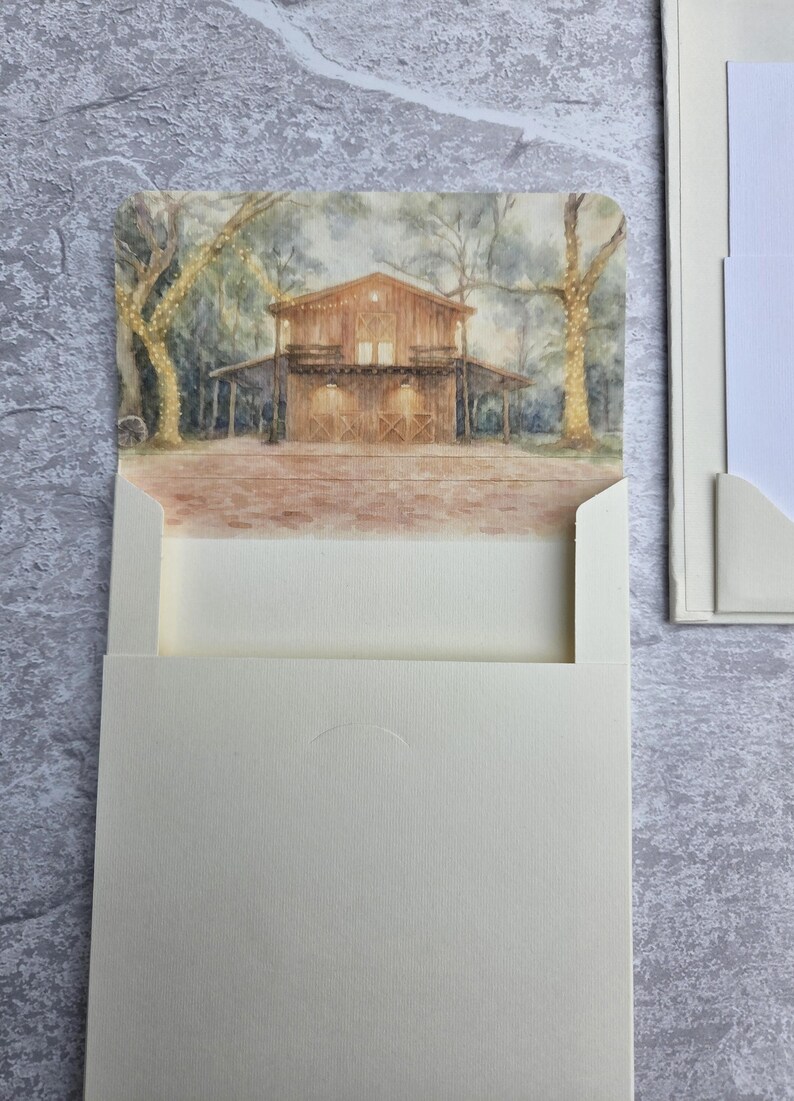 K&ouml;nnte beinhalten: Eine cremefarbene Einladungskarte mit einem Aquarellgem&auml;lde einer rustikalen Scheune mit Lichterketten. Die Karte wird in einen cremefarbenen Umschlag gesteckt, mit einer passenden Karte auf der rechten Seite. Der Hintergrund ist eine strukturierte, hellgraue Oberfl&auml;che.