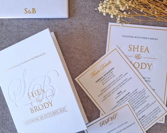 Könnte beinhalten: Weiße Hochzeitseinladung mit goldener Schrift. Die Einladung lautet "Sie sind herzlich eingeladen zur Hochzeit von Shea & Brody, Samstag, 28. Oktober 2023".