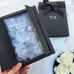 Invito di matrimonio di lusso in scatola nera, con petali bianchi e stampa in foil bianco e nero su acrilico trasparente.