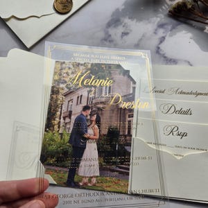 May include: A clear acrylic wedding invitation with gold foil accents. The invitation features a photo of a couple in front of a building. The text on the invitation reads "Because you have shared a special part in their lives, Melanie & Preston".