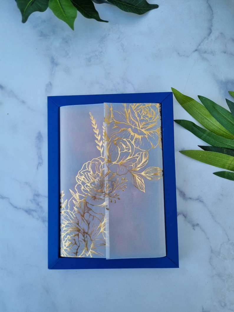 May include: A rectangular blue card holder or frame featuring a translucent vellum overlay. The vellum is decorated with a metallic gold floral line art design, showcasing detailed illustrations of blooming flowers and foliage on the front surface.