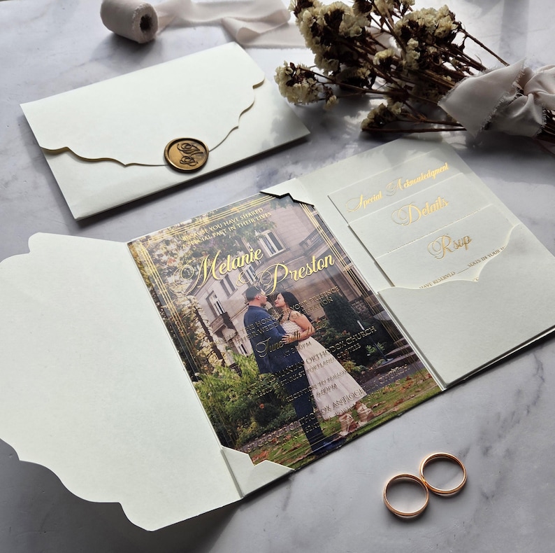 May include: A white wedding invitation with a gold foil design. The invitation features a photo of the couple in front of a building. The invitation also includes RSVP cards and a details card.