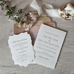 May include: A white wedding invitation suite featuring embossed floral borders and grey text. The main card reads Amy Maree Hollis and Thomas Luca Durand Pucci, Sunday 06 December 2026. A smaller scalloped card titled The Finer Details is included.
