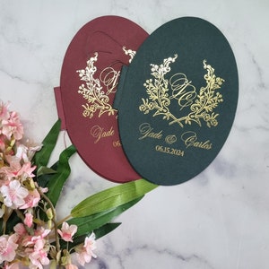 May include: Two oval-shaped wedding invitations with gold foil lettering. The invitations are burgundy and dark green. The text on the invitations reads "Jade & Carlos 06.15.2024".