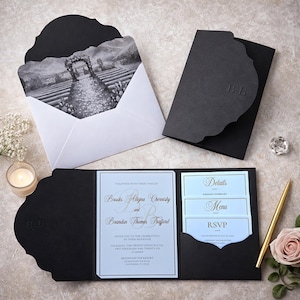 Op de afbeelding: Elegant zwart trouwuitnodigingspakket met een geschulpte rand. De uitnodiging bevat een zwart-wit landschapsafbeelding en details, menu- en RSVP-kaarten. De uitnodiging is open en toont de namen van het bruidspaar en de huwelijksdetails.
