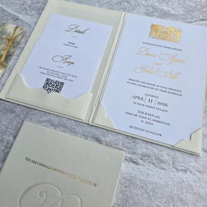 K&ouml;nnte beinhalten: Elfenbein-Hochzeitseinladungs-Suite mit Goldfolientext. Die Einladung enth&auml;lt Details, RSVP und Veranstaltungsinformationen. Die Suite wird in einem Ordner im Taschenstil mit einer passenden Karte pr&auml;sentiert.