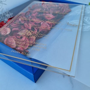 May include: A clear acrylic wedding invitation card featuring gold text and a border, layered over deep red dried flower petals. The invitation details include the names Muhammad Fahad Nasir, the date October 28, 2022, and the World's Fair Marina location.