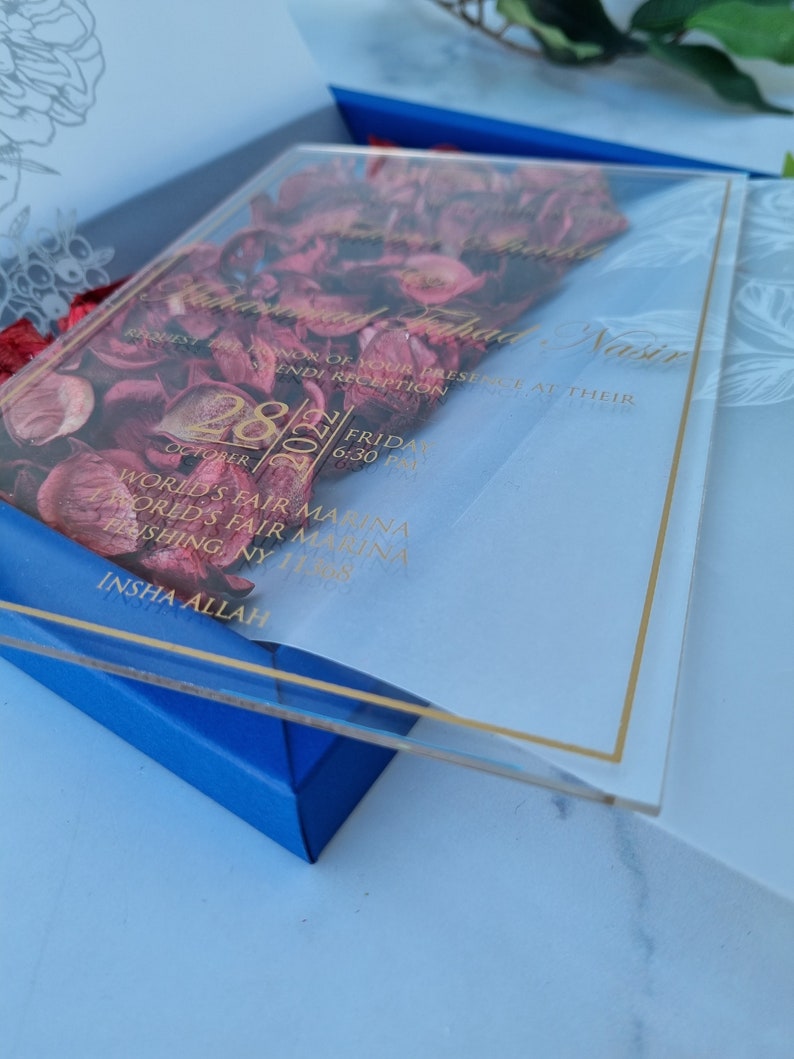 May include: A clear acrylic wedding invitation card featuring gold text and a border, layered over deep red dried flower petals. The invitation details include the names Muhammad Fahad Nasir, the date October 28, 2022, and the World's Fair Marina location.
