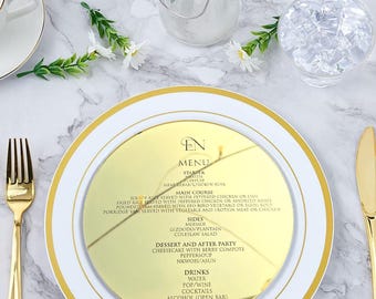 May include: A gold-rimmed white plate with a gold menu insert, fork, and knife. The menu lists starters, main courses, sides, dessert, and drinks. The menu is on a marble surface with a cup, water glass, and floral accents.