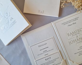 Könnte beinhalten: Eine weiße Hochzeitseinladung mit einem silbernen Prägedesign. Die Einladung enthält die Namen "Ramiro Colmenero" und "Johanna Valiente" sowie das Datum "24. Juni 2023". Die Einladung enthält auch Details zur Hochzeitszeremonie und zum Empfang, einschließlich Uhrzeit, Ort und Kleiderordnung.