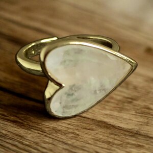 Puede incluir: Un anillo de oro con una piedra preciosa blanca en forma de coraz&oacute;n. El anillo est&aacute; sobre una superficie de madera.