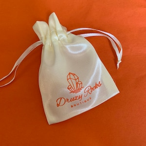 Puede incluir: Una bolsa de cord&oacute;n de sat&eacute;n blanco con un logotipo rosa que dice "Druzy Rocks Boutique" y un gr&aacute;fico de cristal estilizado.