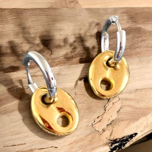 Könnte beinhalten: Ein Paar goldene und silberne Creolen. Die Creolen sind silberfarben und die Anhänger sind goldfarben und oval mit einem kleinen Loch in der Mitte.