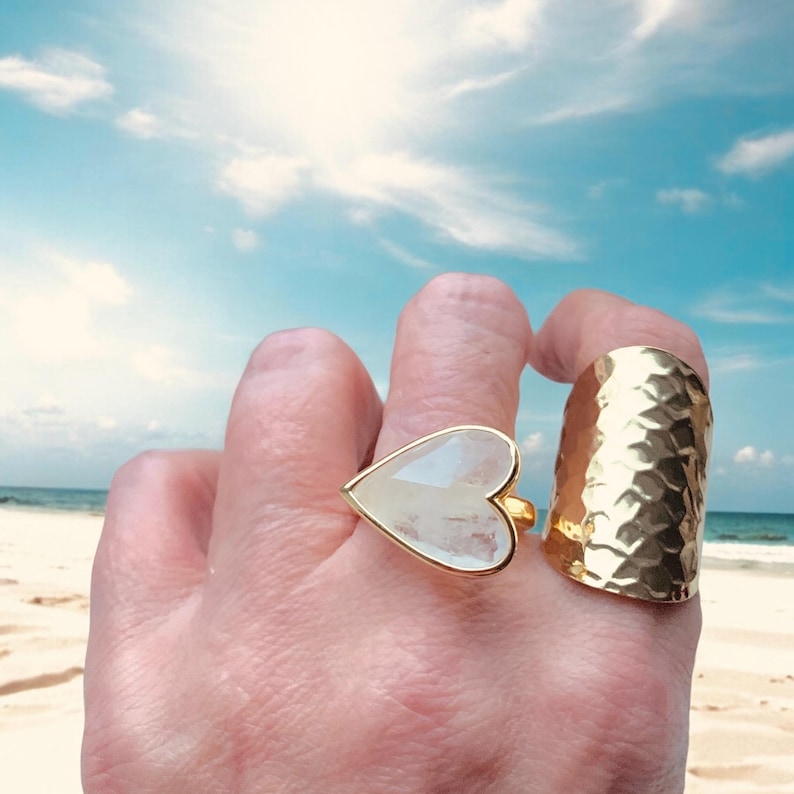 Puede incluir: Dos anillos de oro en una mano. Un anillo es una banda ancha con una textura martillada. El otro anillo presenta una piedra grande, blanca en forma de coraz&oacute;n.