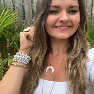 Pode incluir: Uma mulher vestindo uma blusa branca com um colar de corrente dourada com um pingente de lua crescente branca e um pingente retangular branco com detalhes dourados. Ela também está usando uma pilha de pulseiras de contas brancas e douradas no pulso.